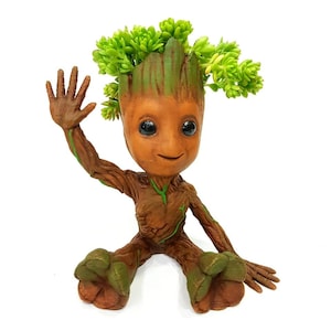 May include: A brown and green figurine of a character from the Guardians of the Galaxy movie, Groot, with a green leafy plant growing out of its head. The figurine is sitting with its right arm raised in a wave.