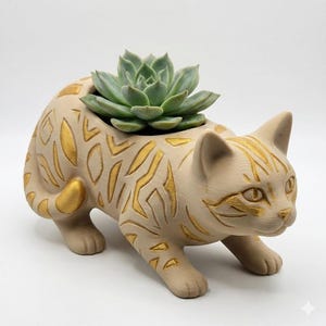 Puede incluir: Una maceta beige con forma de gato con detalles dorados, que contiene una suculenta verde. La maceta de cerámica presenta un diseño detallado, incluyendo patrones grabados y una cara de gato. La suculenta es de color verde claro.