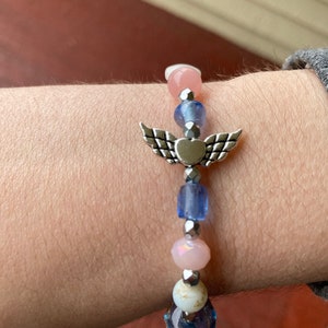 May include: A bracelet with a silver angel wing charm with a heart in the center. The bracelet is made with pink, blue, and white beads.