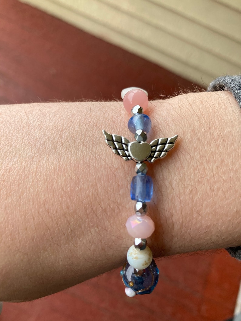 May include: A bracelet with a silver angel wing charm with a heart in the center. The bracelet is made with pink, blue, and white beads.