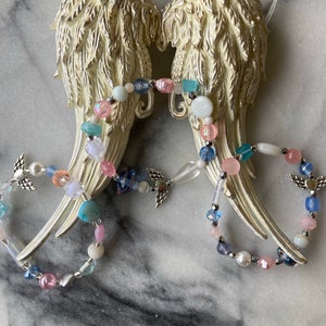 May include: White ceramic angel wings with a beaded garland of pink, blue, and white beads. The garland features small silver angel wings charms.