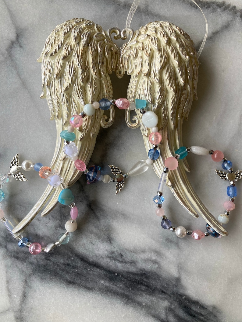 May include: White ceramic angel wings with a beaded garland of pink, blue, and white beads. The garland features small silver angel wings charms.