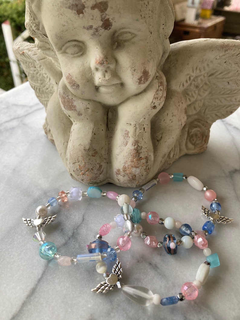 May include: Three beaded bracelets with silver angel wing charms. The bracelets are made with light blue, pink, and white beads.