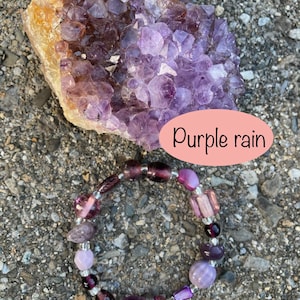 May include: Purple amethyst crystal cluster with a purple and silver beaded bracelet. The bracelet has a variety of purple and clear beads. The text "Purple rain" is written on a pink oval.