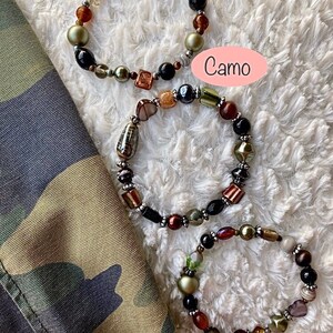 Puede incluir: Tres pulseras de cuentas con una mezcla de cuentas marrones, negras, verdes y doradas. Las pulseras están sobre una superficie blanca y peluda con la palabra "Camo" escrita sobre un fondo rosa.