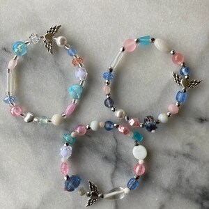 May include: Three beaded bracelets with angel wing charms. The bracelets are made with pink, blue, and white beads.