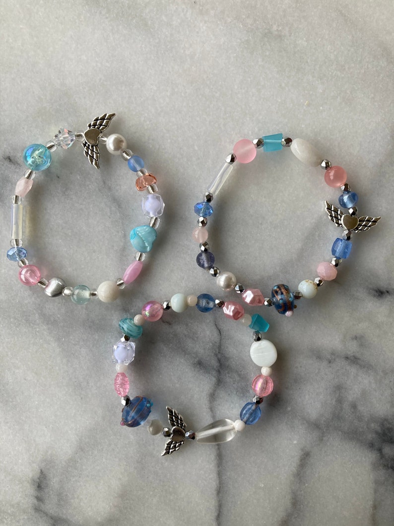 May include: Three beaded bracelets with angel wing charms. The bracelets are made with pink, blue, and white beads.