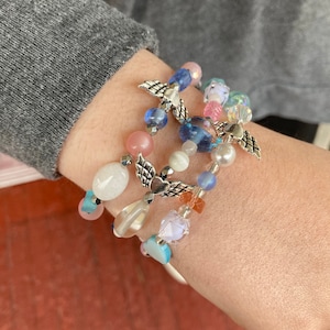 May include: A set of four bracelets featuring colorful beads, including pink, blue, white, and clear glass beads. Each bracelet has a silver charm in the shape of angel wings with a heart in the center.