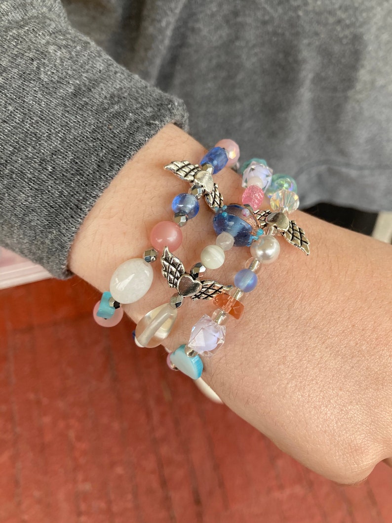 May include: A set of four bracelets featuring colorful beads, including pink, blue, white, and clear glass beads. Each bracelet has a silver charm in the shape of angel wings with a heart in the center.