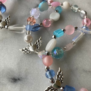 May include: A multi-strand beaded bracelet with silver angel wing charms. The beads are in shades of pink, blue, and white.