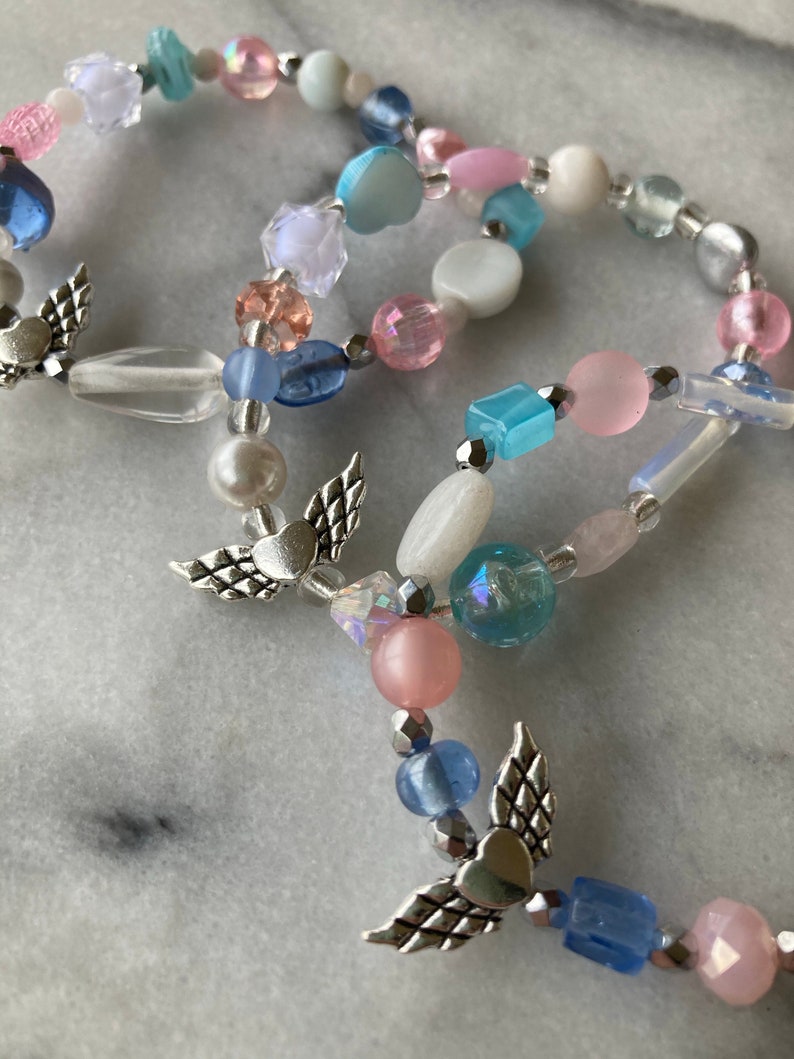 May include: A multi-strand beaded bracelet with silver angel wing charms. The beads are in shades of pink, blue, and white.