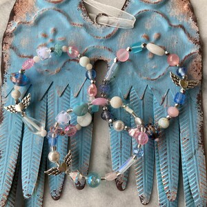 May include: Two beaded angel wing ornaments with silver angel charms, pink, blue, and white beads, and a white ribbon hanging from the top.