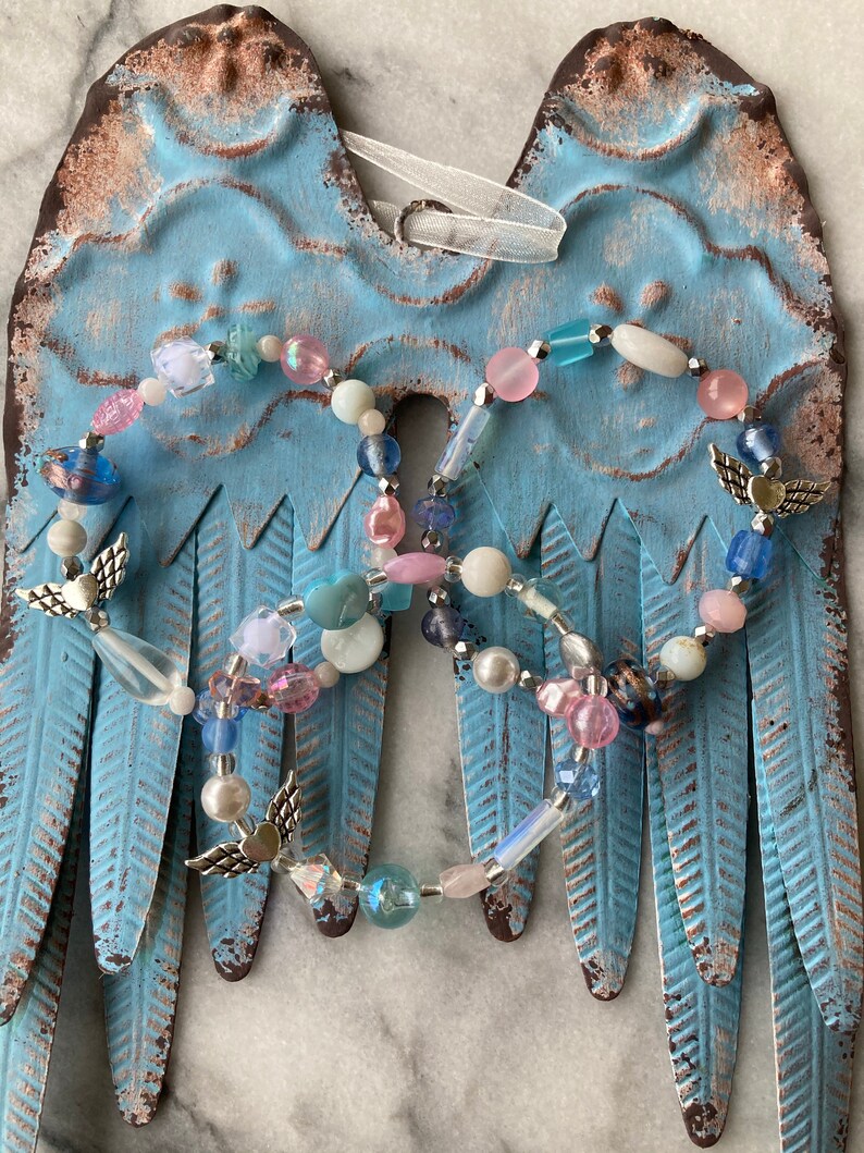 May include: Two beaded angel wing ornaments with silver angel charms, pink, blue, and white beads, and a white ribbon hanging from the top.