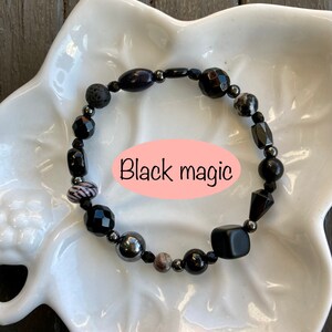 May include: Black beaded bracelet with black lava rock, black onyx, hematite, and other black stones. The bracelet is on a white plate and the text "Black magic" is written on the plate.