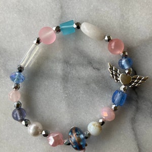 May include: A beaded bracelet with a silver charm of an angel with wings and a heart. The beads are various shades of blue, pink, and white.