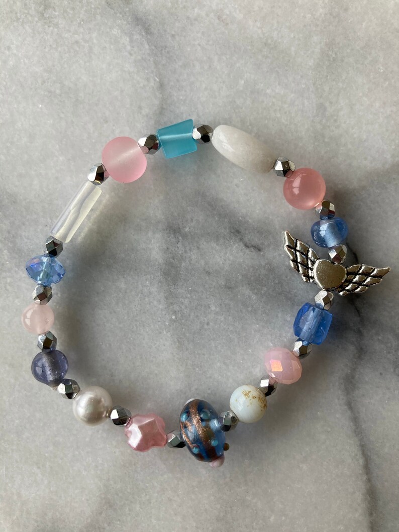 May include: A beaded bracelet with a silver charm of an angel with wings and a heart. The beads are various shades of blue, pink, and white.