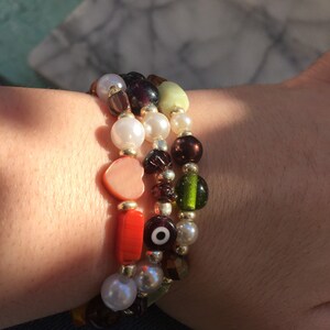 May include: A set of three bracelets with colorful beads, including a heart charm, an evil eye charm, and a variety of other beads in green, red, brown, and white.