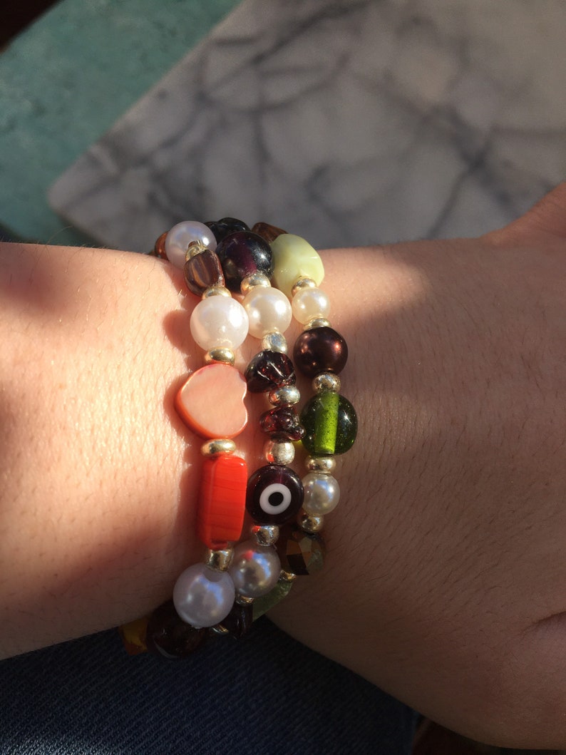 May include: A set of three bracelets with colorful beads, including a heart charm, an evil eye charm, and a variety of other beads in green, red, brown, and white.