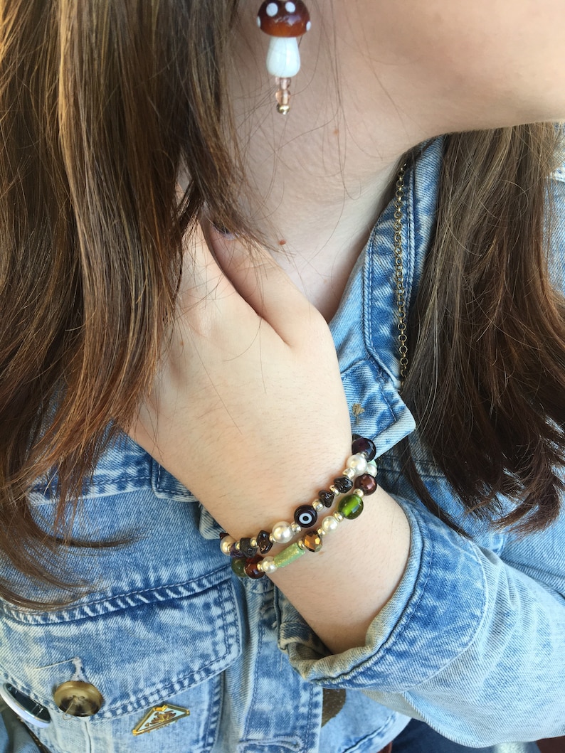 May include: A woman wearing a denim jacket with a mushroom-shaped earring and two beaded bracelets on her wrist. The bracelets are made of green, brown, and white beads.