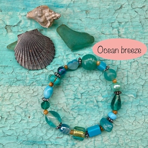 May include: A turquoise blue bracelet with various colored beads, including turquoise, blue, green, and gold. The bracelet is on a turquoise blue background with seashells and sea glass. The text "Ocean breeze" is written in pink on a white background.