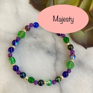 May include: A purple, green, and blue beaded bracelet with silver accents. The bracelet is on a white marble surface with a pink oval label that says "Majesty".