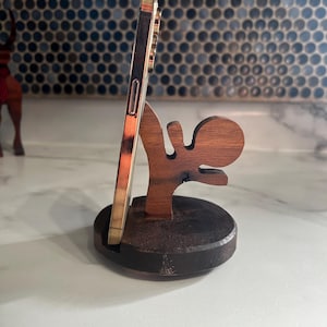 May include: A wooden phone stand in the shape of a person kicking a ball. The stand is made of dark brown wood and has a natural wood finish. A gold smartphone is propped up in the stand.