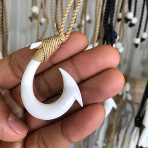 Puede incluir: Colgante de anzuelo de pescado de hueso blanco en un collar de cordón marrón. El anzuelo es un diseño tradicional hawaiano.