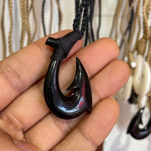 May include: A black wooden fish hook pendant on a black cord necklace. The hook is carved in a traditional style and has a shiny finish.