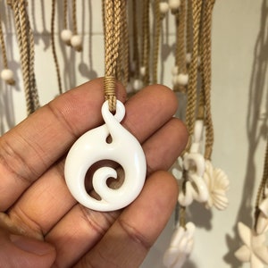 Pode incluir: Um colar com um pingente de osso branco com um design em espiral. O pingente está pendurado em um cordão marrom.