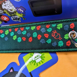 Puede incluir: Un llavero de tela verde con un patrón repetido de fantasmas rojos, verdes y azules. El llavero tiene un anillo de metal plateado.