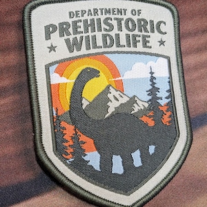 May include: Embroidered patch with the text "DEPARTMENT OF PREHISTORIC WILDLIFE". The patch features a dinosaur silhouette, mountains, trees, and a colorful sunset. The patch has a gray border and is on a brown background.