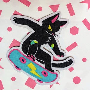 Puede incluir: Gato negro haciendo skateboard en una tabla azul y rosa con un rayo amarillo. El gato tiene una venda en su pierna.