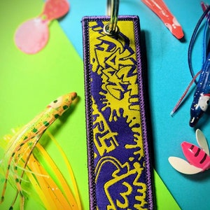 Może przedstawiać: Brelok do kluczy w kolorze fioletowym i żółtym z graficznym wzorem stylizowanej fali. Brelok jest wykonany z tkaniny i ma metalowy pierścień.