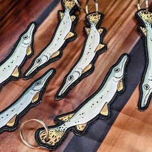 May include: Five keychains featuring a detailed illustration of a longnose gar fish. The fish are depicted in a light gray and white color scheme with black outlines. Each keychain has a gold-colored ring.