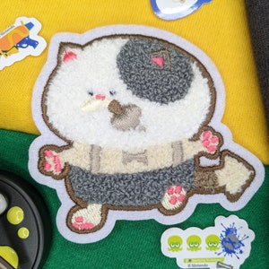 May include: An embroidered patch featuring a white and grey cat wearing a brown waistcoat and a bow tie. The cat has a black patch over one eye and is smiling. The patch is surrounded by a white border.