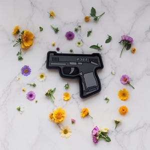 May include: A black and gray pistol-shaped patch with the text "P365" is surrounded by colorful flowers. The flowers are yellow, purple, white, and orange, scattered on a white marble surface. The patch has a detailed design.