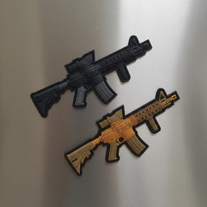 May include: Two toy rifles are displayed. One is black with gray accents, and the other is tan with brown accents. Both have detailed designs, including scopes and magazines. The rifles are likely made of plastic or a similar material.