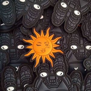 以下が含まれることがあります： 黒とオレンジのワッペンがいくつか集まっているアップ写真。ワッペンには、人間の顔をしたスタイリッシュな太陽と双眼鏡が描かれている。