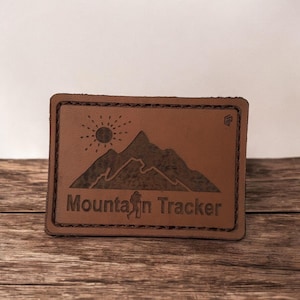 Puede incluir: Un parche rectangular de cuero marrón con las palabras "Mountain Tracker" y un diseño de cordillera. El parche también presenta un sol y una silueta de excursionista. Los bordes están cosidos con hilo marrón más oscuro.
