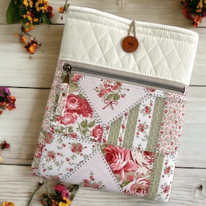May include: Quilted fabric tablet sleeve with a floral patchwork design. The top section is cream-colored with a diamond pattern and a wooden button closure. The main body features a mix of floral and striped patterns in shades of pink, red, and green.