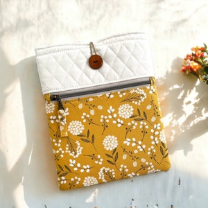 May include: A mustard yellow and white floral pattern tablet sleeve with a white quilted top section. The sleeve has a gray zipper and a brown button closure. The design features white floral patterns on a mustard yellow background.