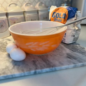 Könnte beinhalten: Nahaufnahme einer weißen Keramik-Arbeitsplatte mit einer Schüssel, einem Schneebesen, zwei Eiern und einer Tüte Gold Medal Mehl. Die Schüssel ist orange mit einem weißen Weizenmuster. Der Schneebesen ist silberfarben und die Eier sind weiß.