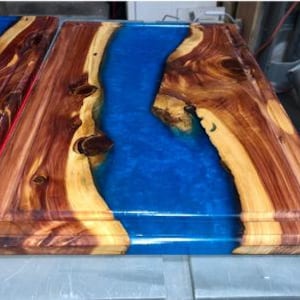 May include: A wooden cutting board with a blue epoxy resin inlay. The board has a natural wood grain and a raised edge.