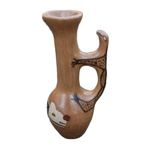 May include: A brown ceramic vase with a unique handle and a detailed design. The vase features a white and brown animal face on the base and a black and brown animal figure on the handle. The vase has a narrow neck and a wide base.