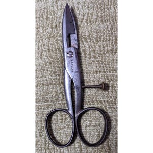 May include: Vintage metal scissors with a pointed blade and looped finger holes. The scissors have a screw adjustment and the name "B. BARCLAY" is visible on the side. The scissors are set against a textured, light-colored background.