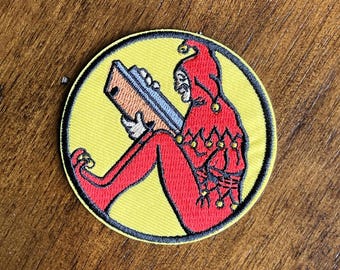 May include: A red and yellow embroidered patch of a jester reading a book. The jester is wearing a red and yellow hat with bells and a red and yellow costume with bells.