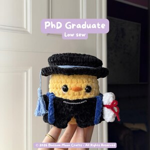 May include: A handmade, crocheted chick wearing a graduation cap and gown. The chick is yellow with black eyes and an orange beak. The cap is black with a blue tassel, and the gown is black with blue trim. The chick holds a white scroll tied with red ribbon. Text reads "PhD Graduate Low sew".