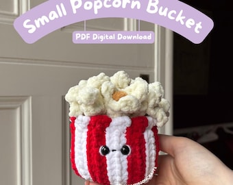May include: A crocheted small popcorn bucket with red and white vertical stripes, featuring a face with two black eyes and a small mouth. The top is filled with textured white popcorn pieces. Text reads Small Popcorn Bucket and PDF Digital Download.