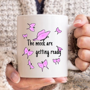 May include: White ceramic mug with a pink butterfly design and the text "The meek are getting ready".