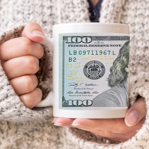 Puede incluir: Taza de cerámica blanca con un diseño de un billete de 100 $ con Benjamin Franklin. El billete tiene el número de serie LB 09711967 L y el texto "Federal Reserve Note".